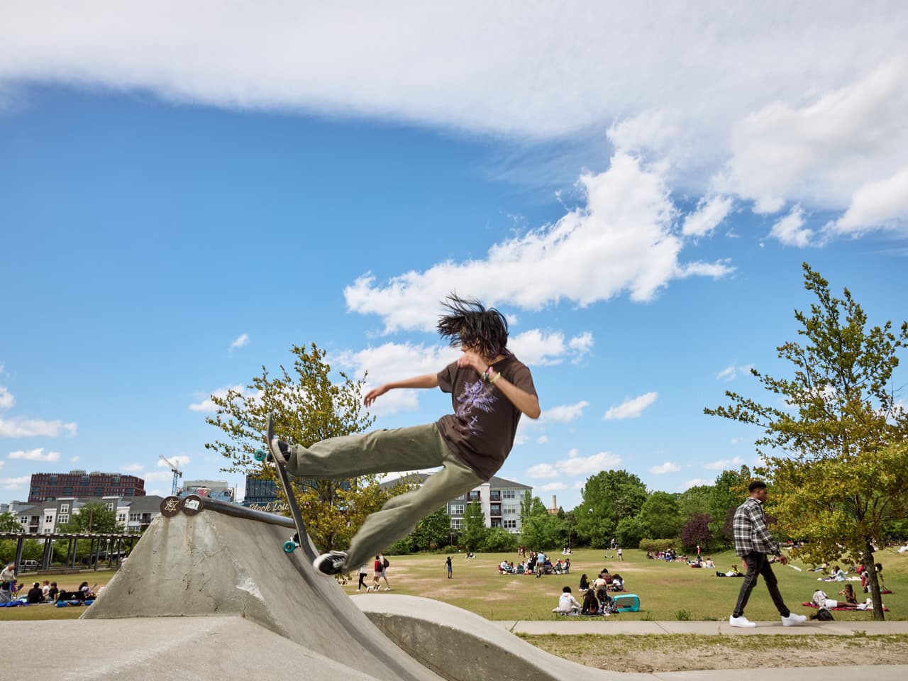 A man rides a skateboard to the top of a ramp, with his hair blowing in the wind. Behind him, people sit in groups on a grass activity field.