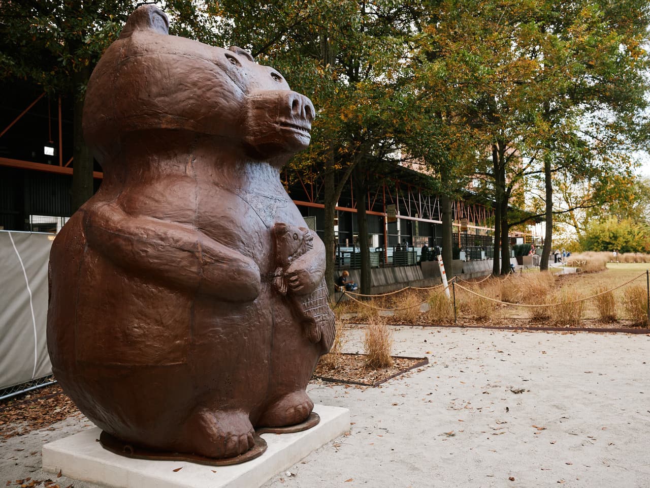 Large, brown bear sculpture holding a smaller fish, set back in the trees, near a modern building.