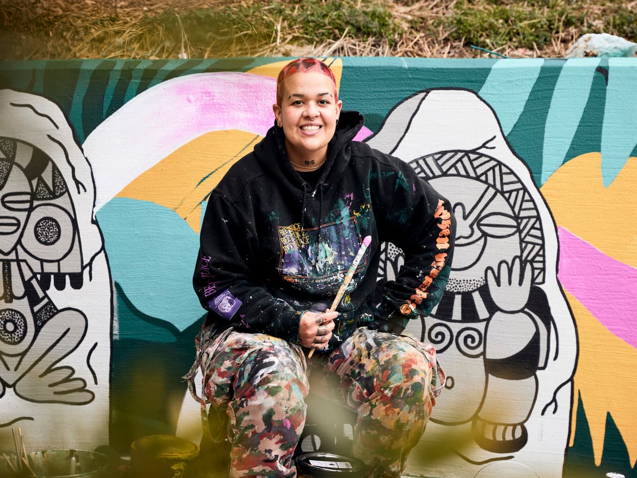 Artist smiling, seated in front of a colorful mural with abstract figures. They hold a paintbrush and wear a paint-splattered outfit.