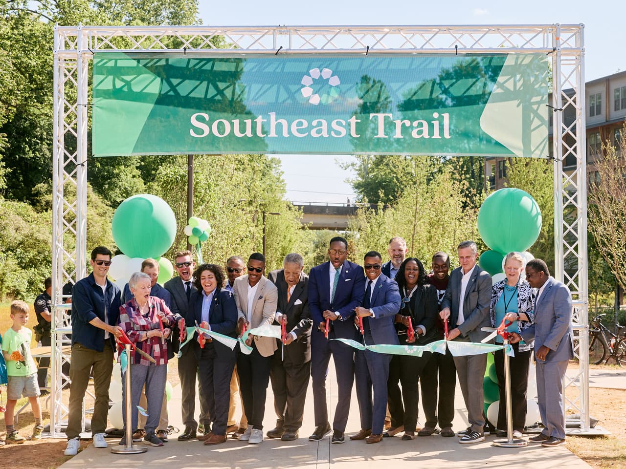 A group of people cut a ribbon under a "Southeast Trail" banner during a sunny outdoor ceremony, surrounded by greenery and balloons.