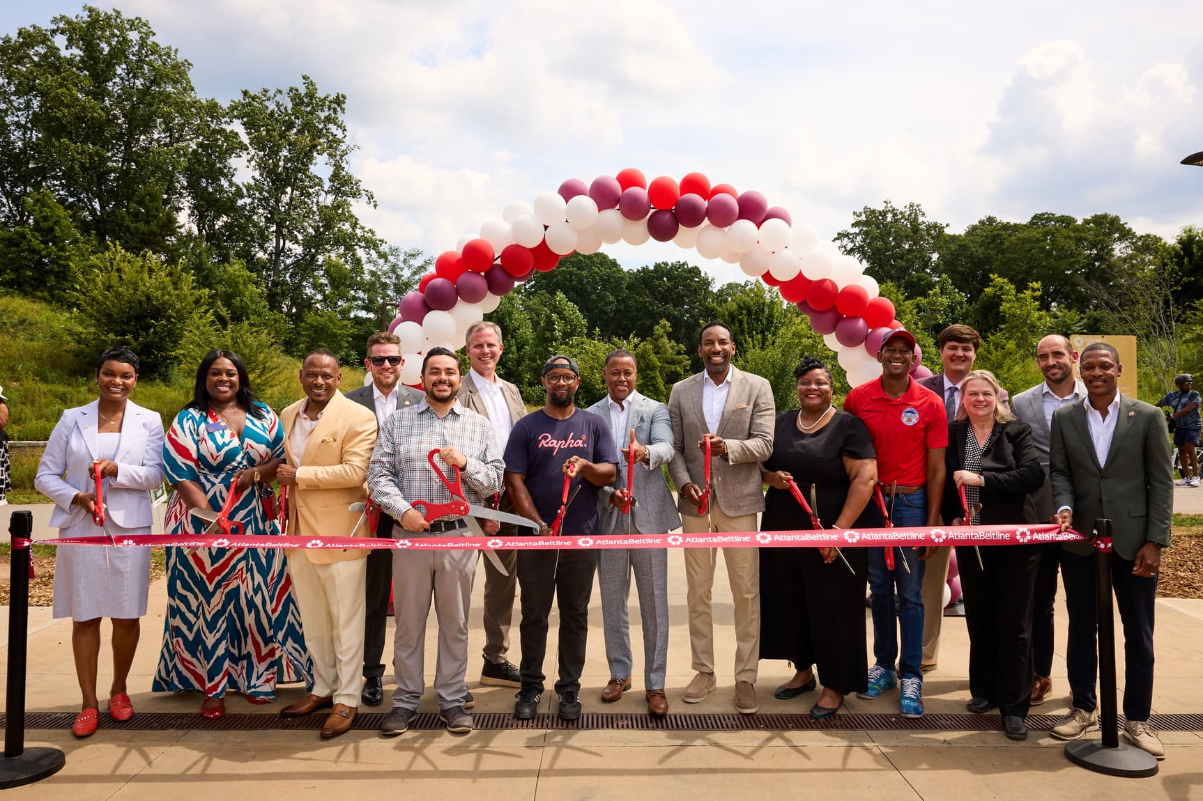 A diverse group of people is cutting a ribbon under a balloon arch at an outdoor event, celebrating a new opening with a backdrop of trees.