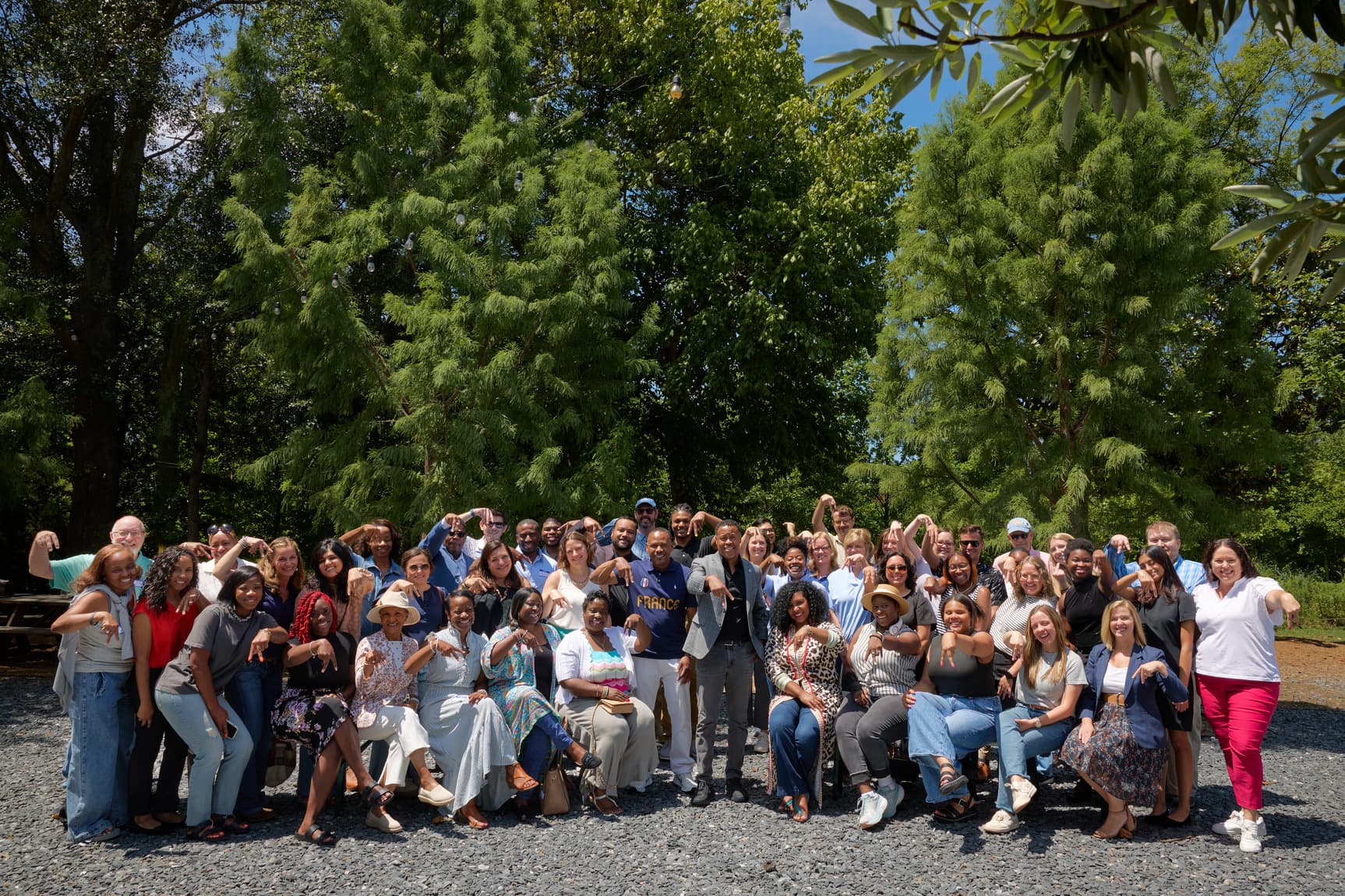 A large group of people posing outdoors, some making playful gestures, with trees in the background and a sunny sky above.