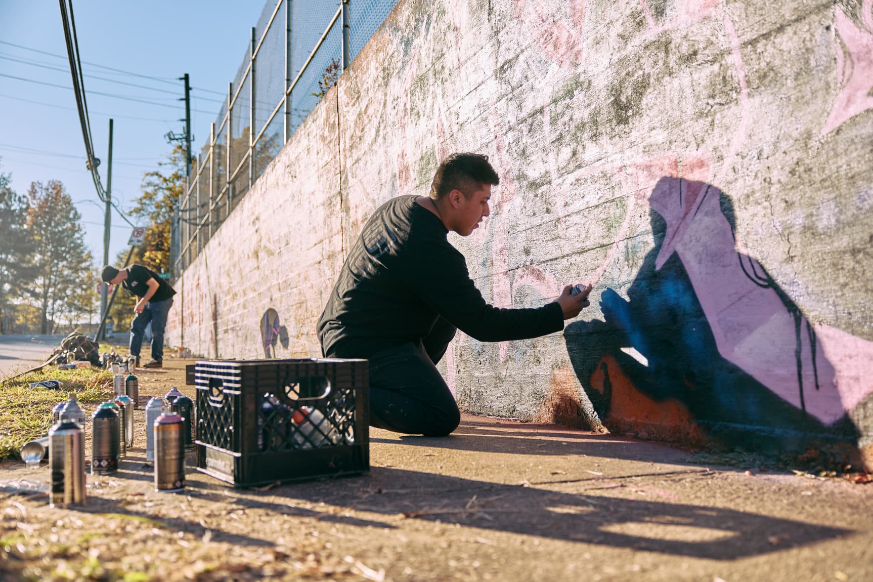Two artists spray-paint a mural on a sunlit wall, surrounded by paint cans. One kneels, focused on details; the other works in the background.