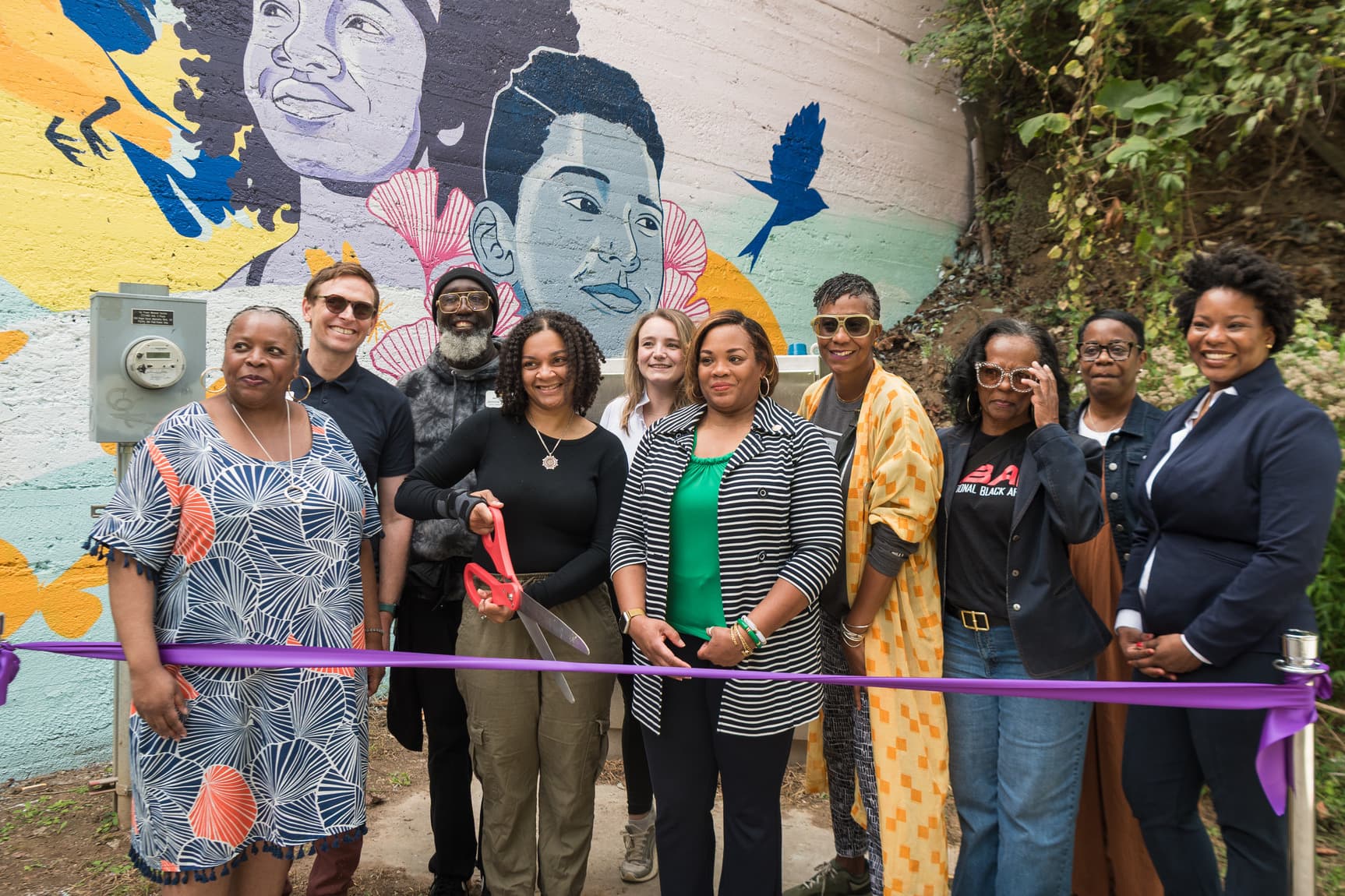 A group of people stand in front of a vibrant mural, holding a ribbon for a cutting ceremony. The mural features colorful portraits and birds.