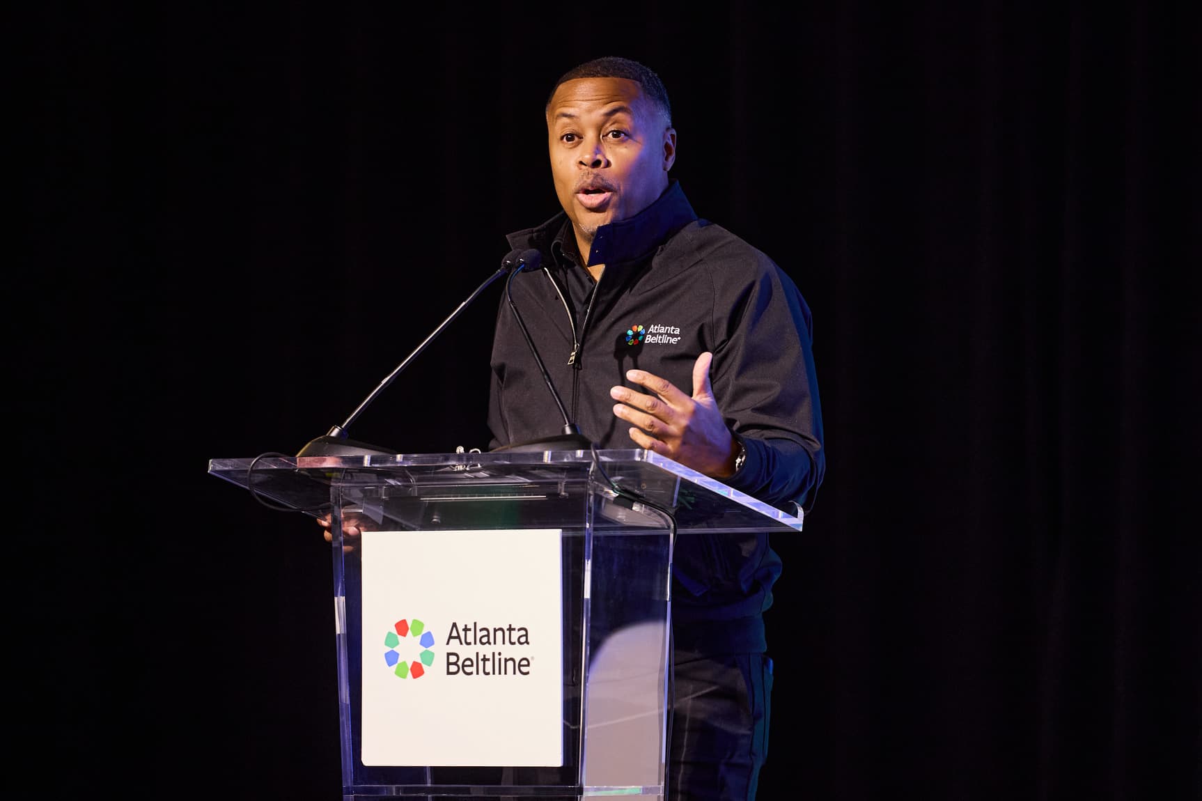 Man speaking at a podium adorned with the Atlanta Beltline logo, wearing a black jacket, with a dark backdrop.
