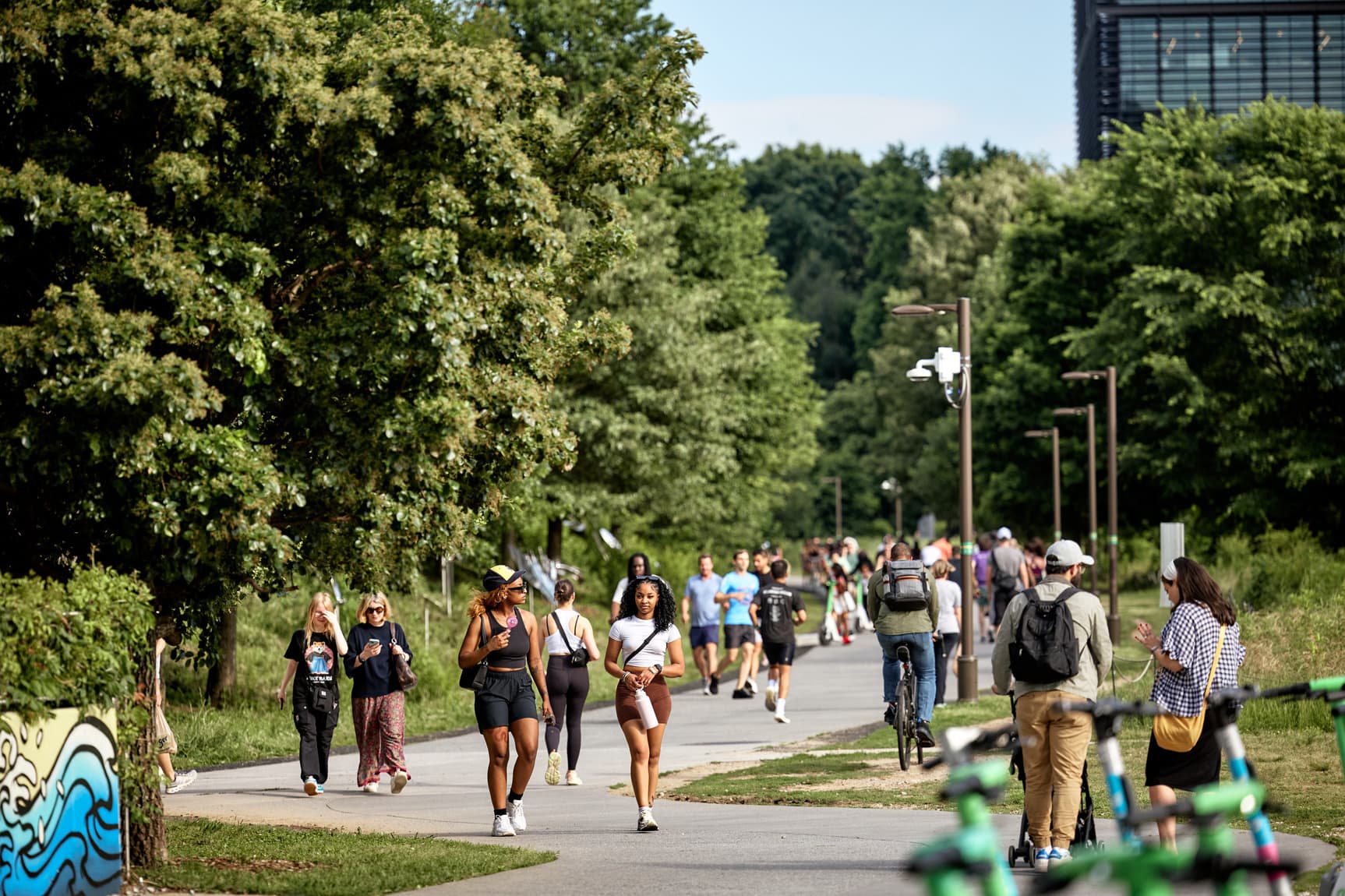 People walking and biking on a tree-lined path, with lush greenery and a building in the background.
