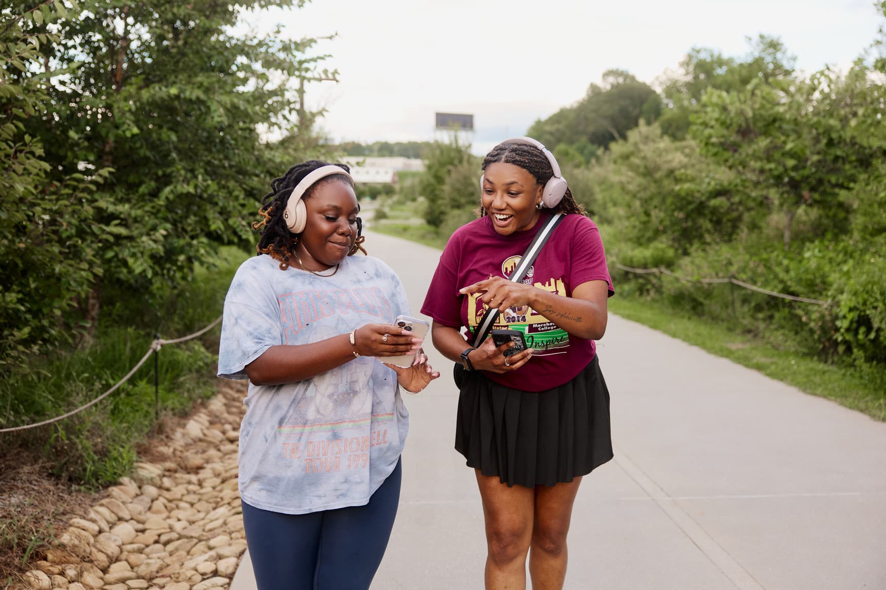 Two people with headphones walk along a path, smiling and looking at a phone, surrounded by greenery.