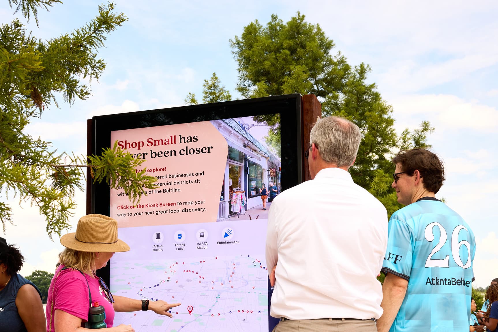 Three people stand at an outdoor kiosk displaying a "Shop Small" message, interacting with a map under a clear sky.