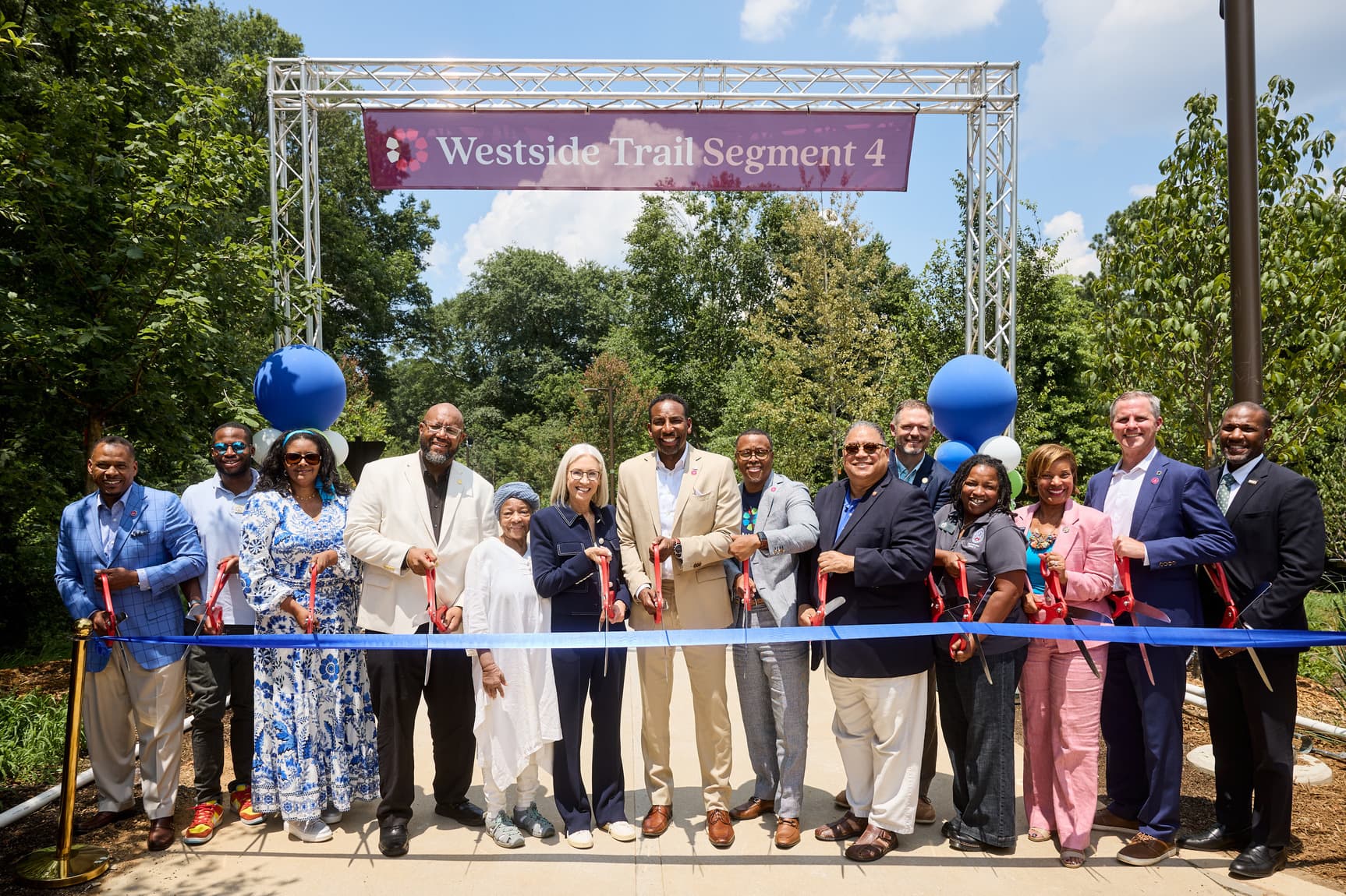 A photo of a group of people posing together, holding giant scissors, and standing behind a big blue ribbon. Behind them is Westside Trail - Segment 4.