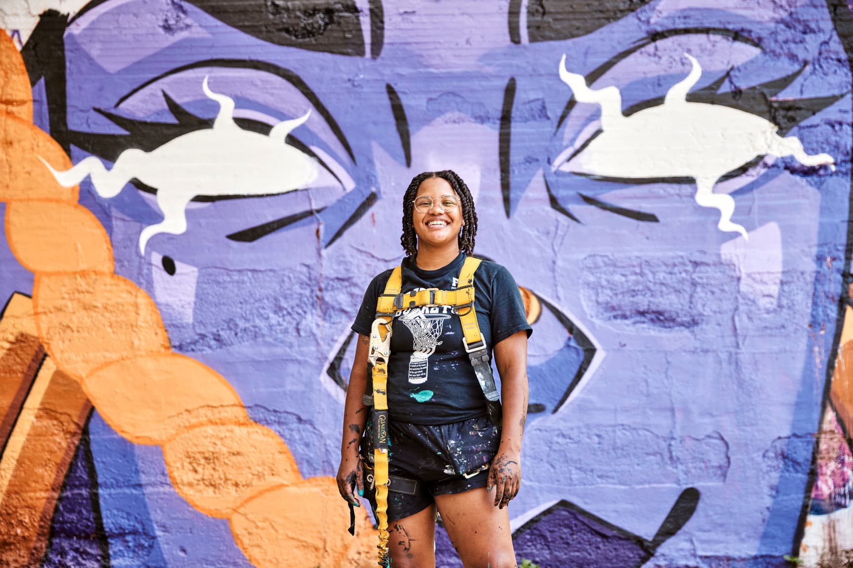 An artists stands, smiling and covered in paint, in front of a colorful mural.
