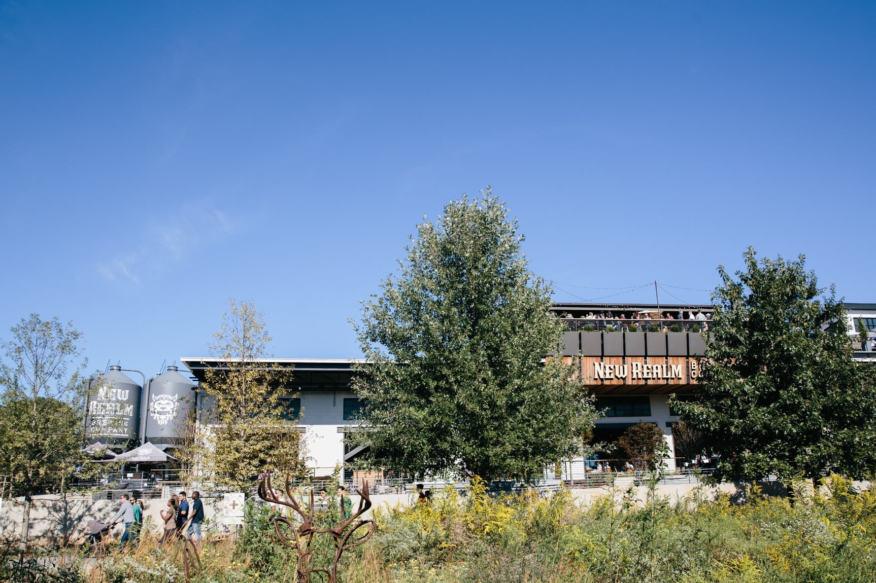 Exterior view of New Realm Brewing Co. with trees and clear blue sky, featuring outdoor seating and large brewing tanks.