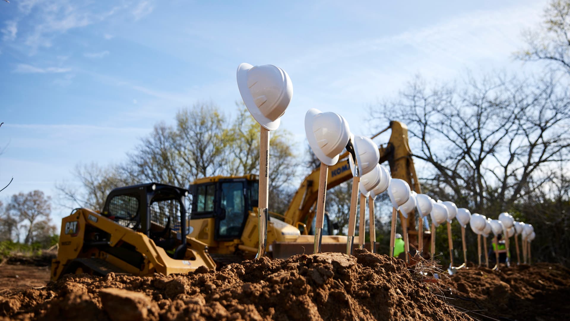 Construction equipment sits behind a line of shovels, standing in dirt, with construction hard hats balanced on top of them.