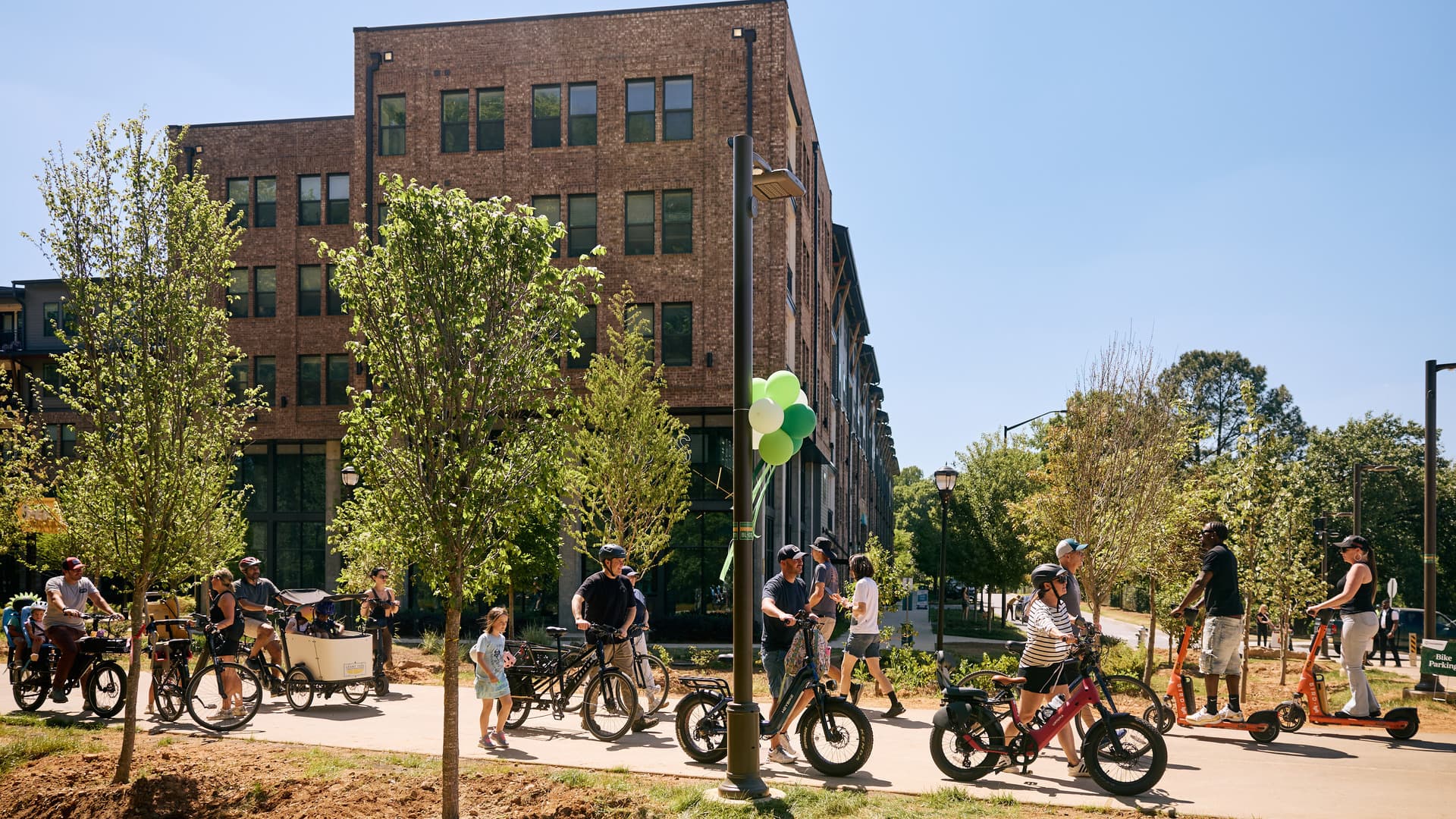 People riding bikes and electric scooters on a sunny path lined with trees and a brick building.