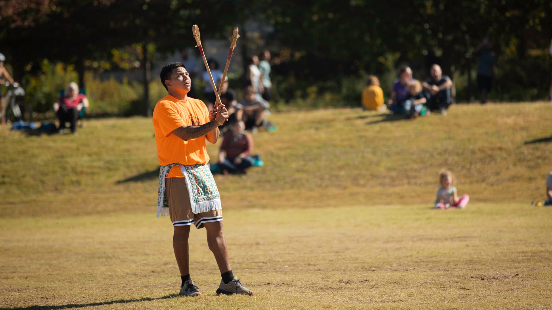 A person stands, holding two toli sticks, with a ball in the air. Nearby, people watch while sitting on a hill.