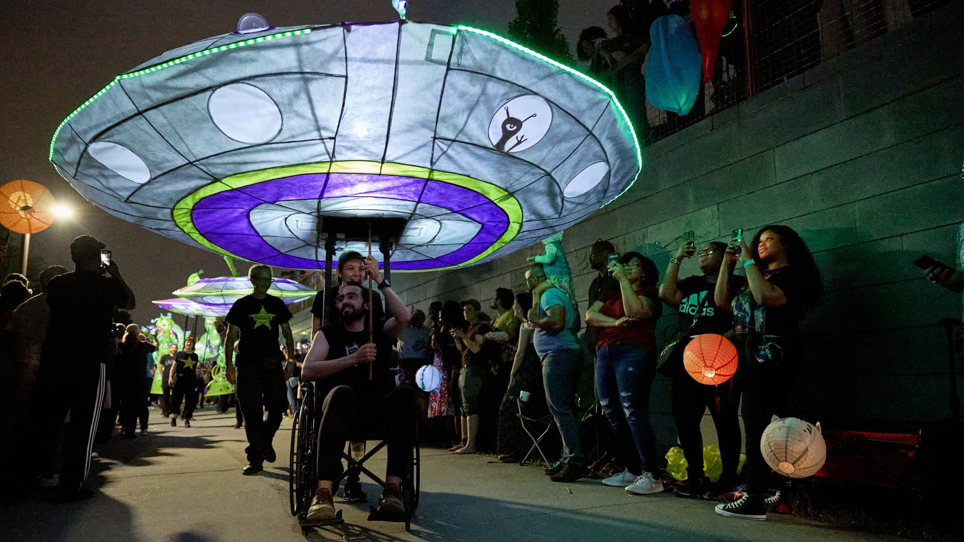 A man in a wheelchair holds a large illuminated puppet of an alien spaceship.
