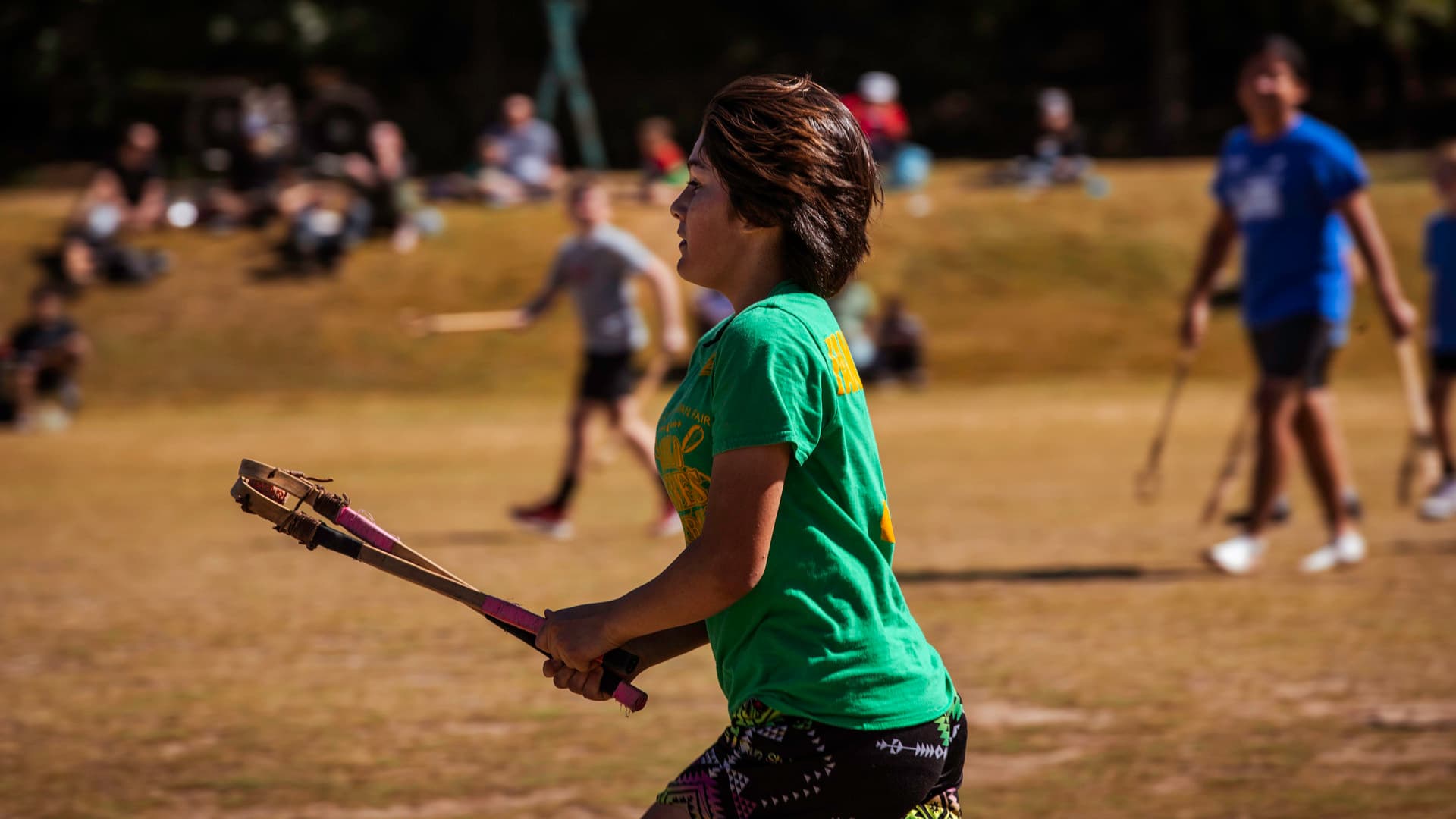 A Stickball player holds their sticks in front of them, while others run on the field in the background.