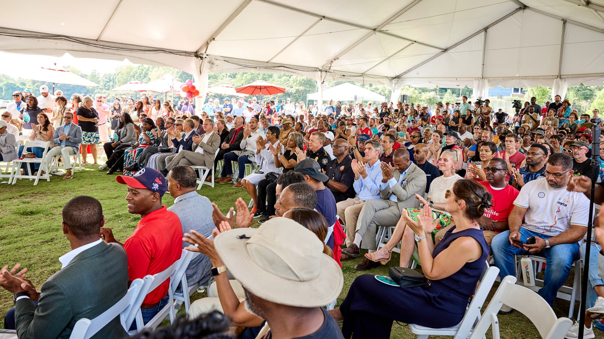 A large, diverse crowd seated under a tent applauding at an outdoor event, with some standing at the back, surrounded by trees and sunshine.