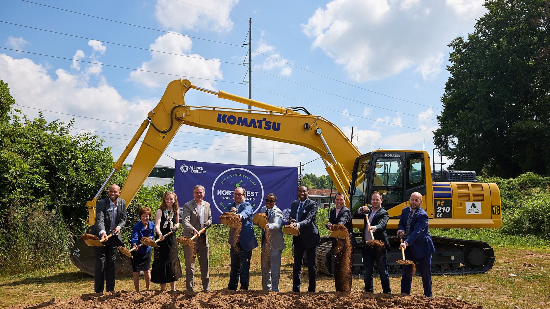 A group of people stand behind a pile of dirt and in front of construction equipment, holding shovels.