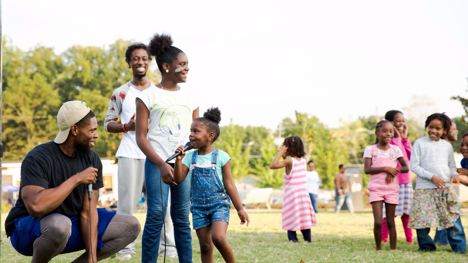 A girl sings into a microphone while others smile around her at a park.
